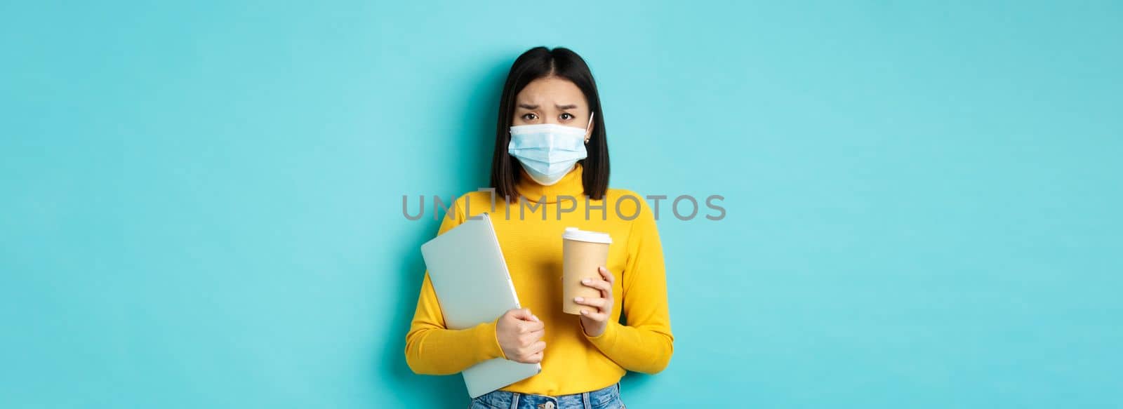 Covid-19, social distancing and pandemic concept. Worried asian woman in medical mask, frowning sad, holding laptop for work and cup of coffee by Benzoix