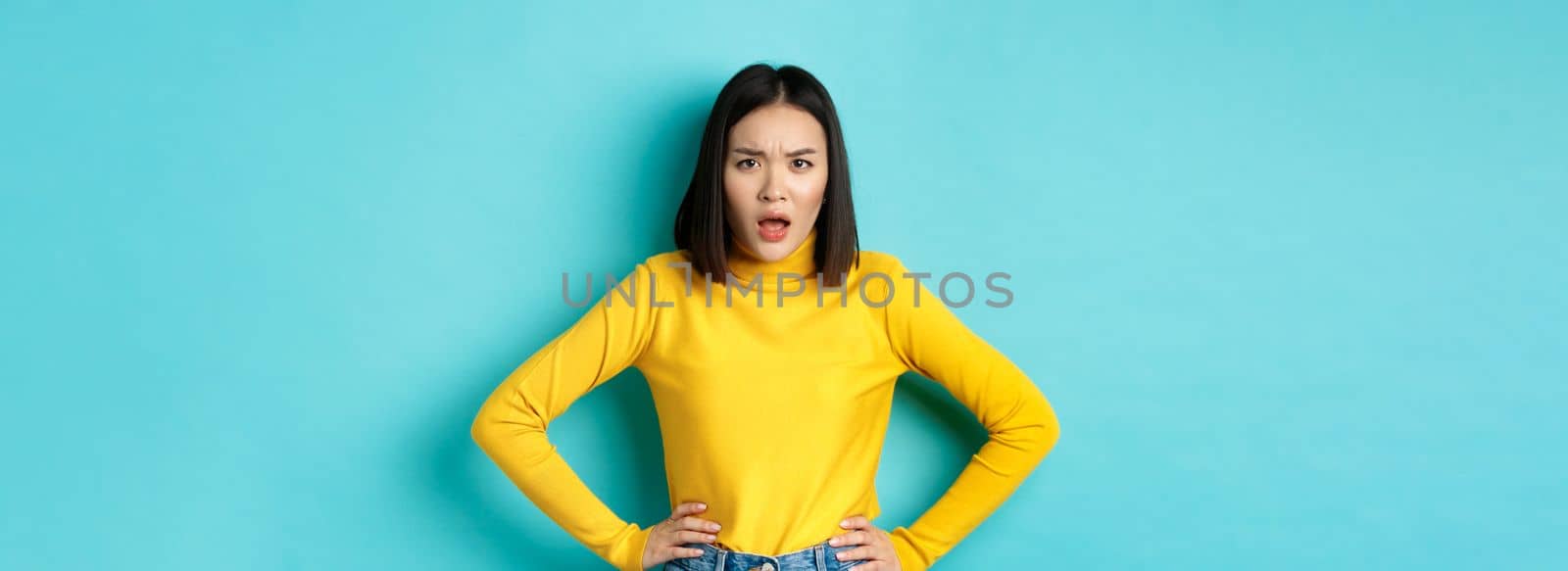 Portrait of angry and confused asian woman staring frustrated at camera, standing against blue background by Benzoix