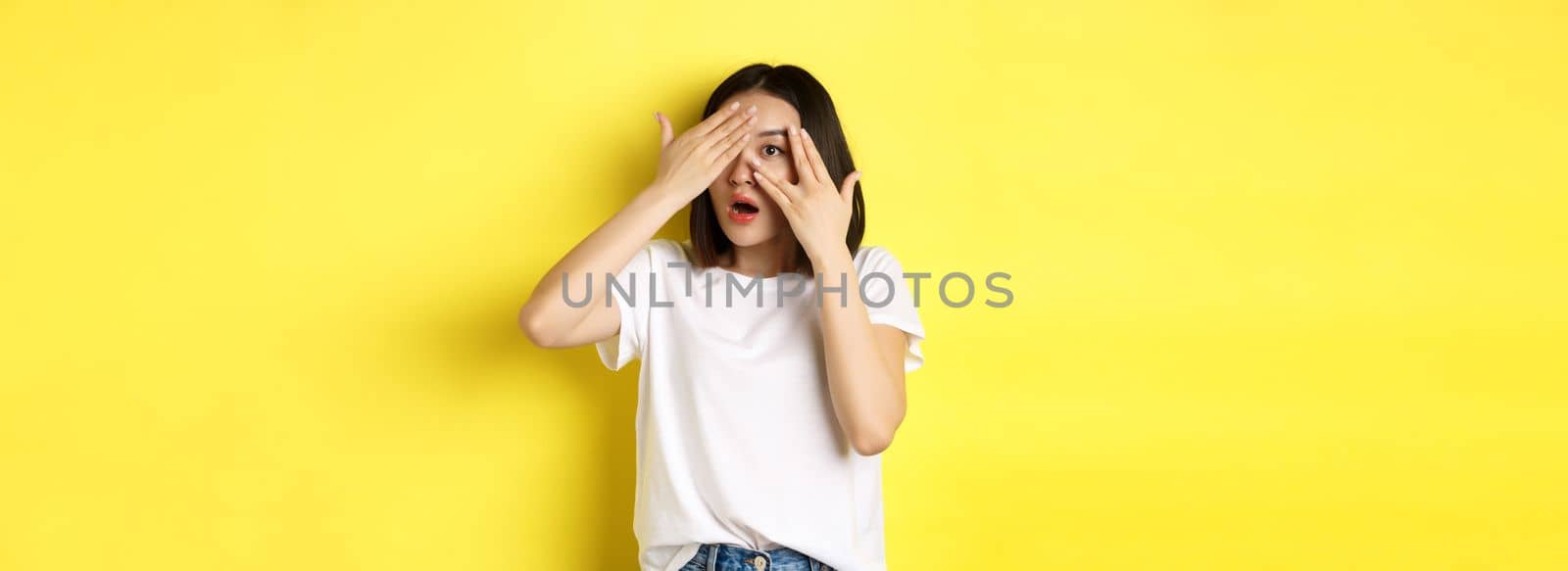Intrigued asian girl waiting for surprise, peeking through fingers on eyes, smiling happy, standing over yellow background.