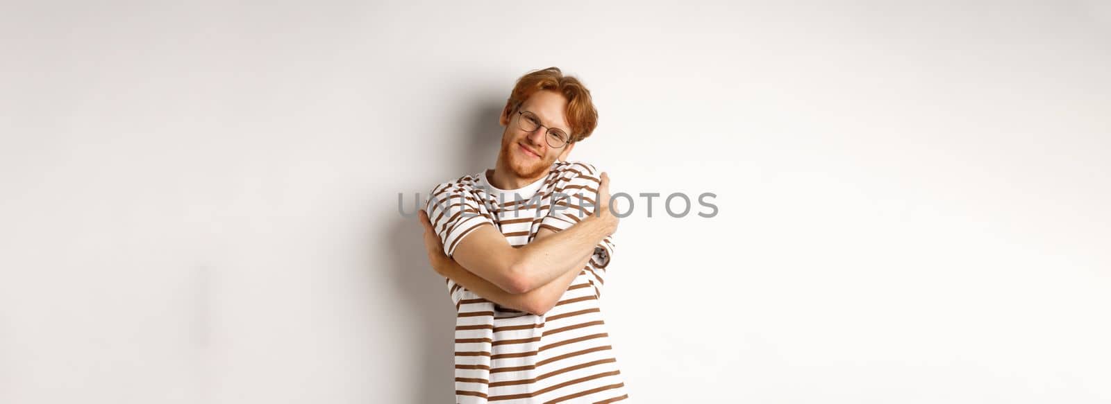 Handsome smiling man with red hair and beard looking relaxed, hugging himself and staring at camera happy face, white background.