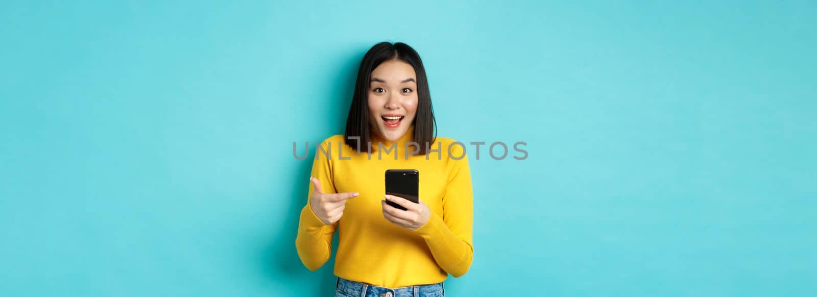 E-commerce and online shopping concept. Surprised asian woman demonstrate smartphone app, internet discounts, pointing finger at mobile phone, blue background by Benzoix