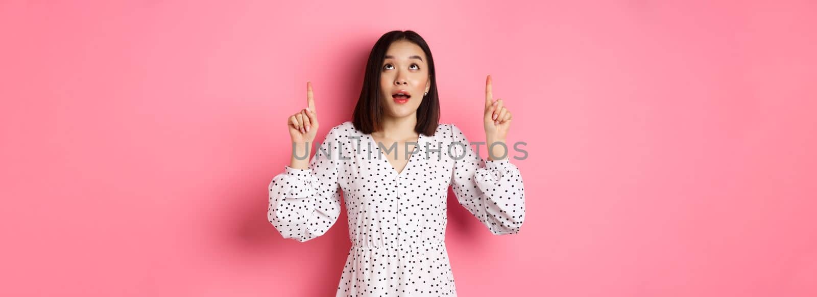 Image of beautiful asian girl in dress looking, pointing fingers up, gazing dreamy at promo offer, standing over pink background by Benzoix