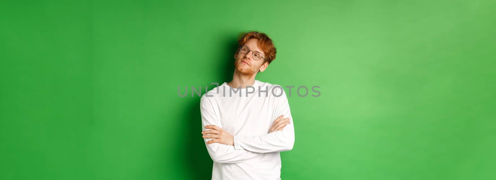 Confident young man with red hair, wearing glasses, looking smug at right side copy space, cross arms on chest self-assured, standing over green background.