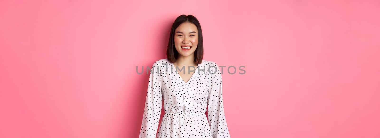 Happy korean woman in dress looking at camera, smiling and laughing with sincere expression, standing over pink background. Copy space