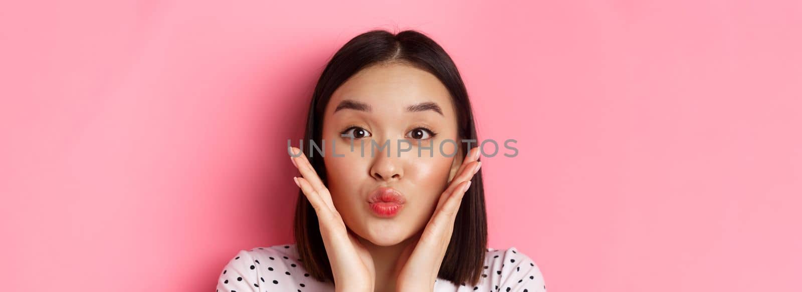 Beauty and lifestyle concept. Close-up of adorable asian woman touching face, pucker lips in kiss, standing over pink background.
