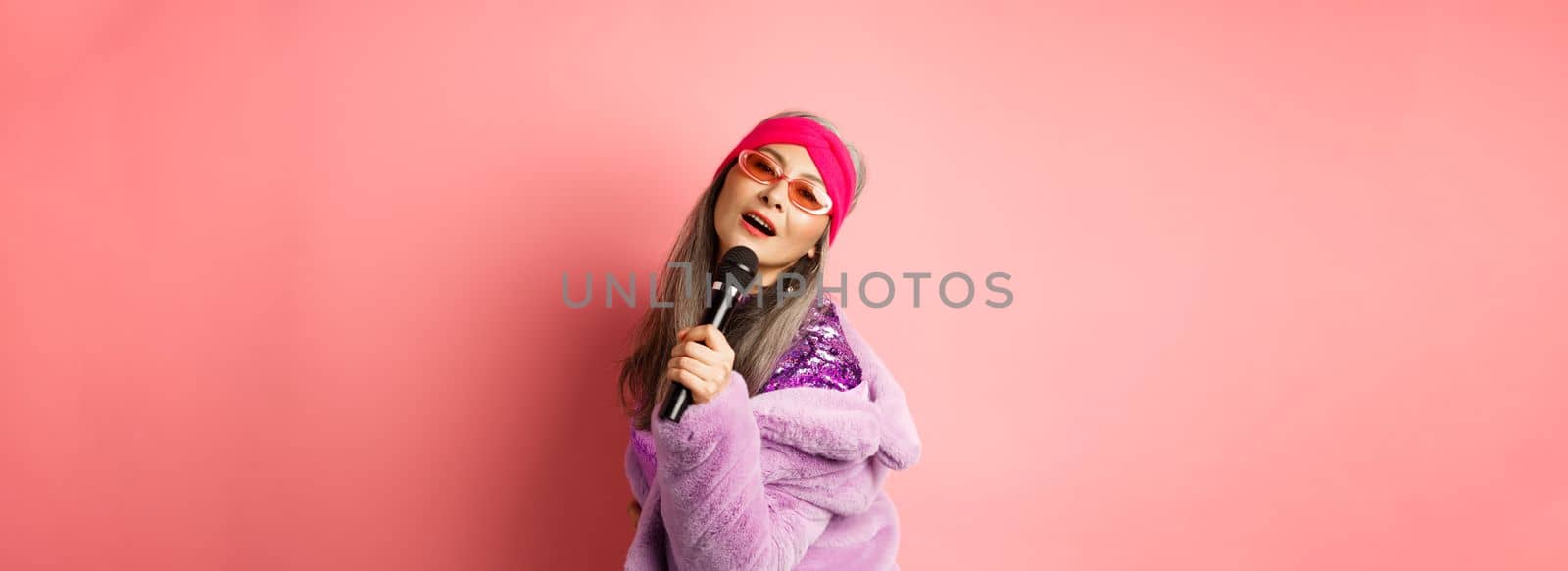 Stylish and sassy asian mature woman perform song on stage, holding mic and singing karaoke, wearing trendy heart-shaped sunglasses and faux fur coat, pink background.