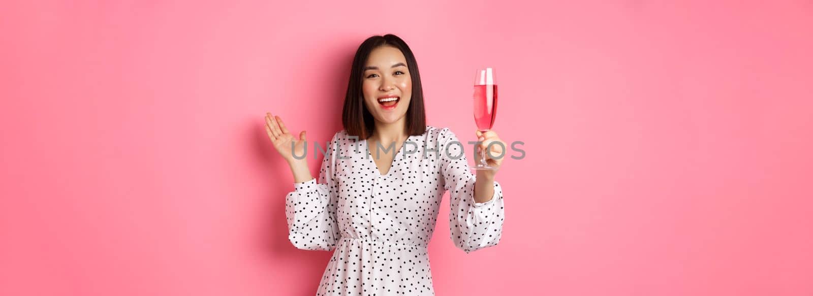 Happy asian woman celebrating, saying toast on party, raising glass of champagne and smiling, standing in dress over pink background by Benzoix