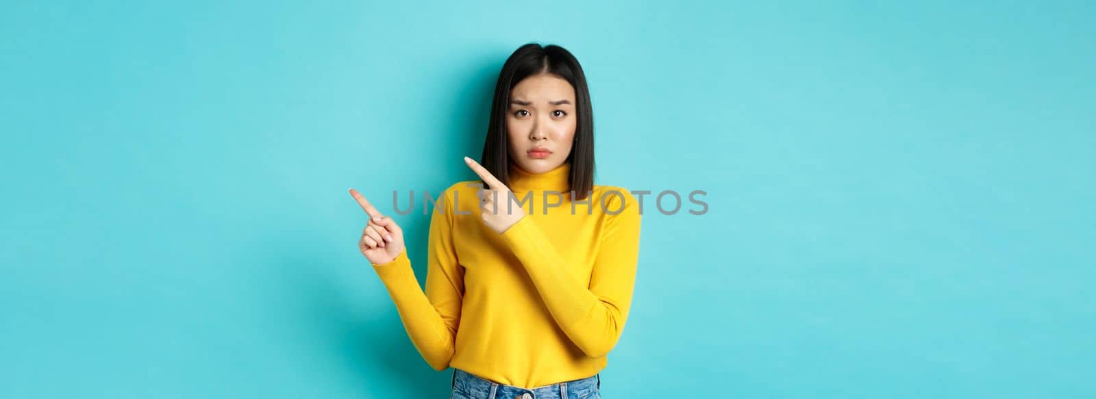 Shopping concept. Disappointed korean girl looking gloomy, asking to buy this, pointing fingers at upper right corner and staring sad at camera, blue background.