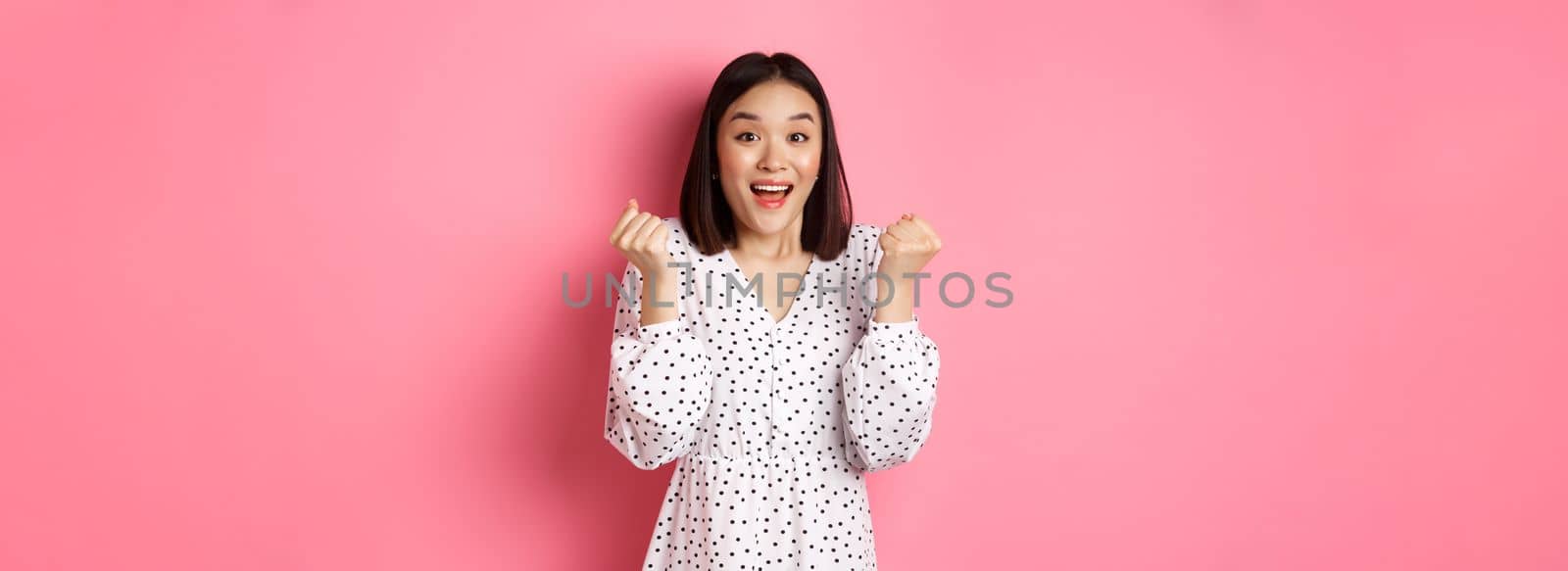 Image of beautiful asian woman winning, clench fists and saying yes with happy face, triumphing and celebrating victory, standing over pink background.
