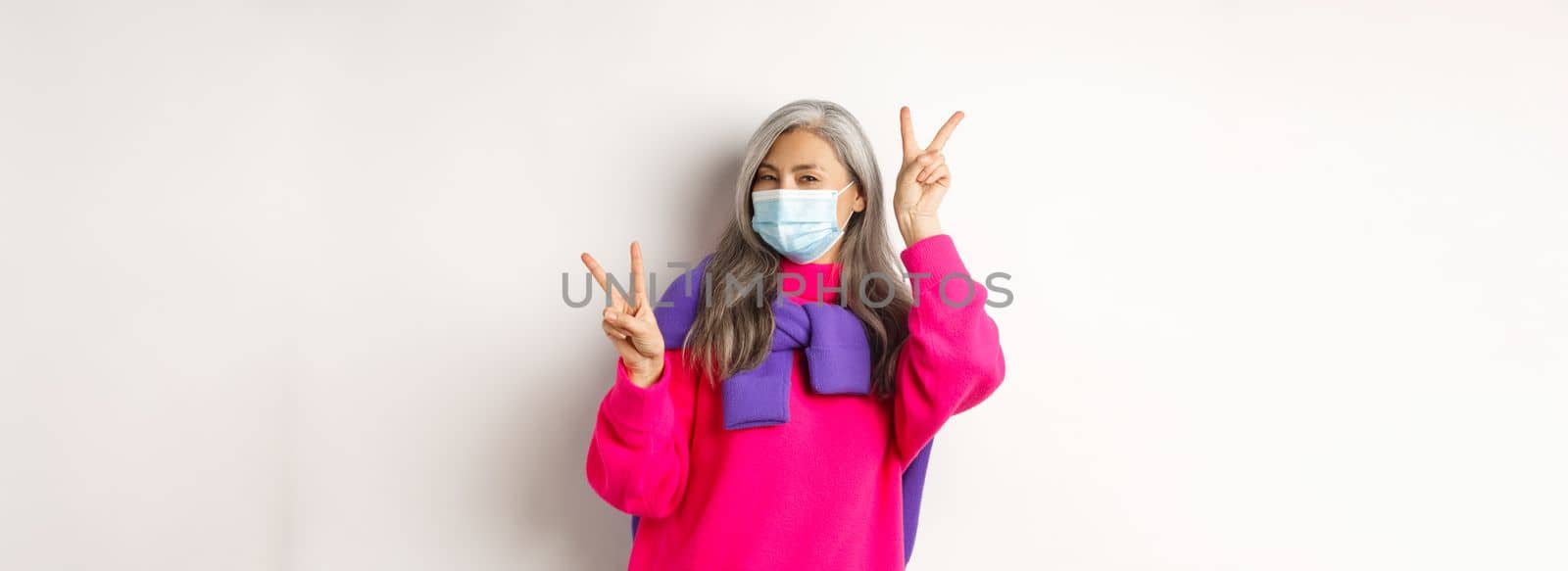Covid, pandemic and social distancing concept. Cheerful and stylish asian senior woman wearing medical mask and showing peace signs, standing over white background.
