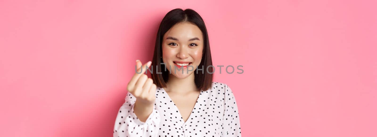 Beauty and lifestyle concept. Close-up of lovely asian woman showing heart sign, smiling and feeling romantic on valentines day, standing over pink background by Benzoix