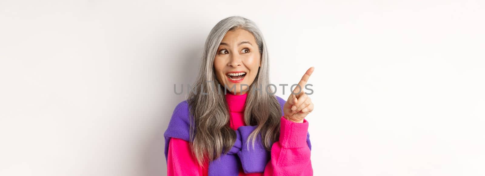 Close-up of stylish korean senior woman with grey hair looking and pointing upper left corner, showing promotion offer, standing over white background.