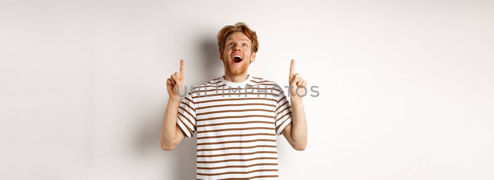 Fascinated redhead man looking with amazement and happiness, pointing fingers up, checking out something cool, standing over white background.