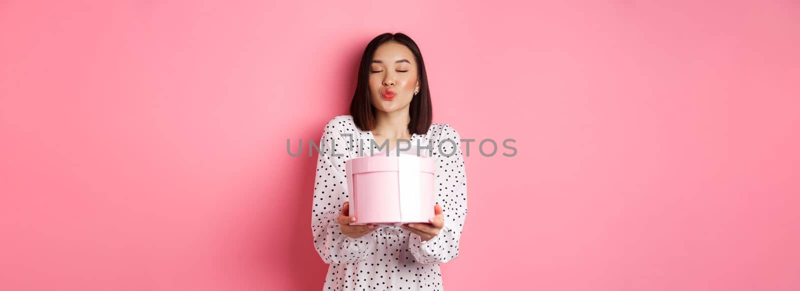 Cute asian girlfriend congratulate with valentines day, giving cute romantic gift in box, pucker lips for kiss, standing over pink background.