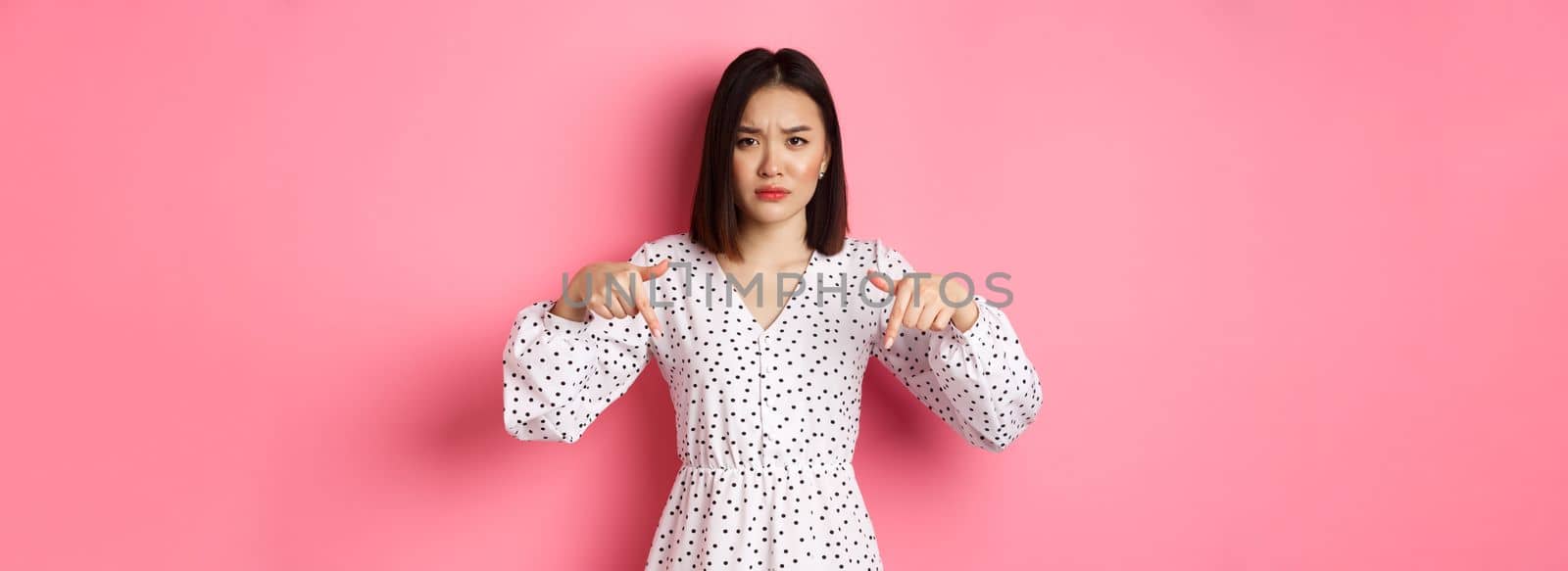 Disappointed asian female model pointing fingers down at promo offer, frowning and staring at camera upset, standing over pink background.