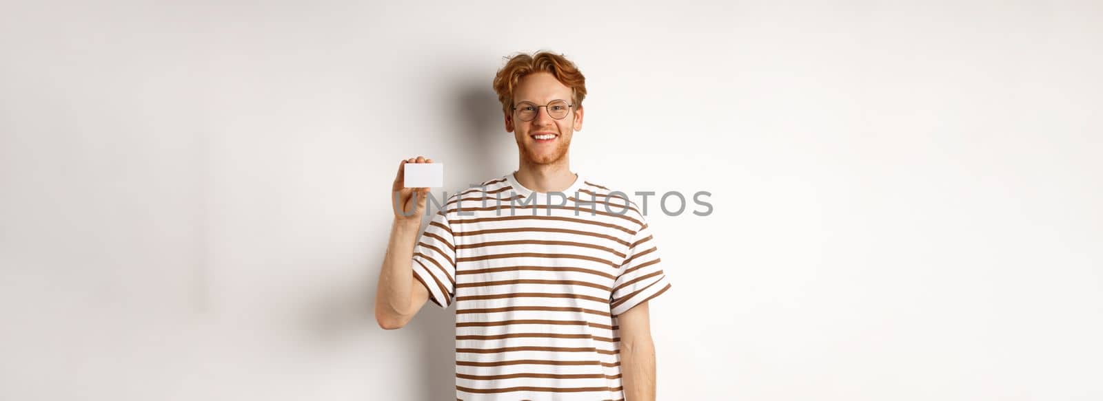 Shopping and finance concept. Smiling young man with bristle and red hair showing plastic credit card and looking happy, white background.
