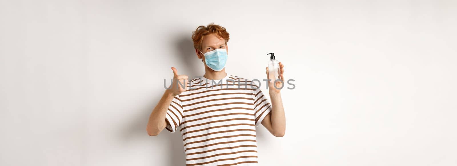 Covid-19, health and lifestyle concept. Smiling male model with red hair, wearing face mask, showing hand sanitizer and thumb-up, recommend to use antiseptic, white background.