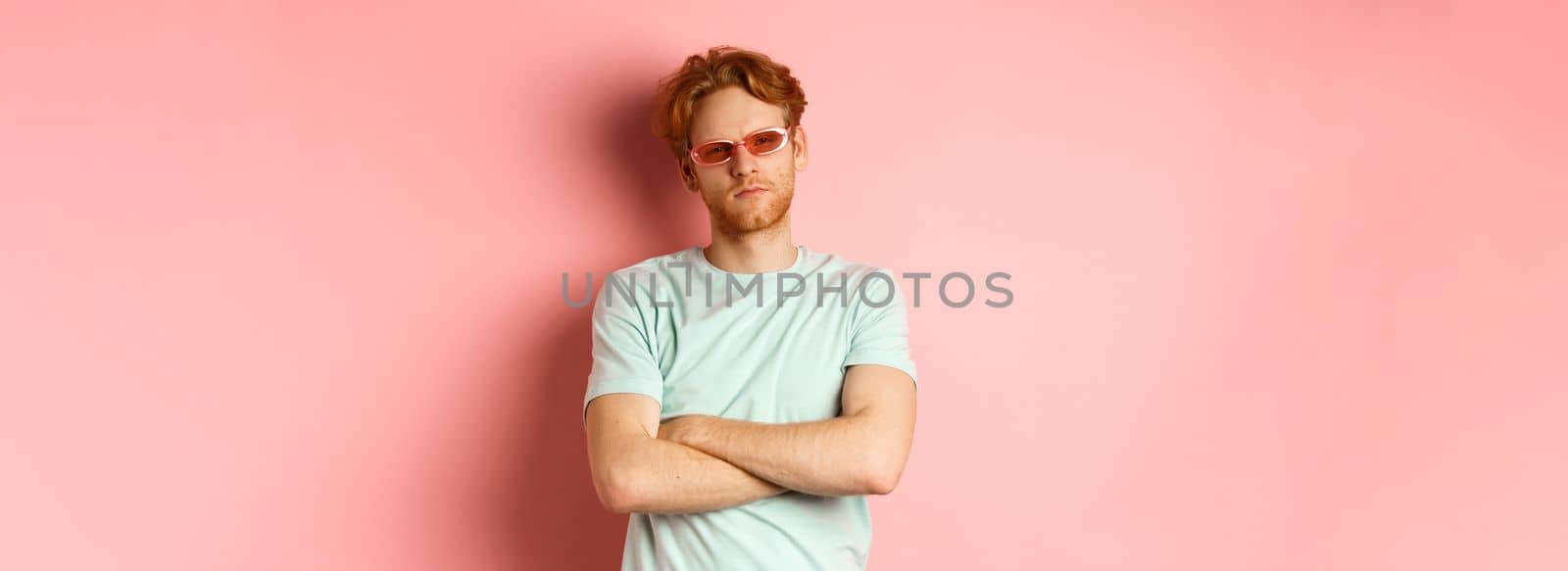 Tourism and vacation concept. Sassy redhead guy in sunglasses, cross arms on chest and looking serious at camera, standing over pink background by Benzoix