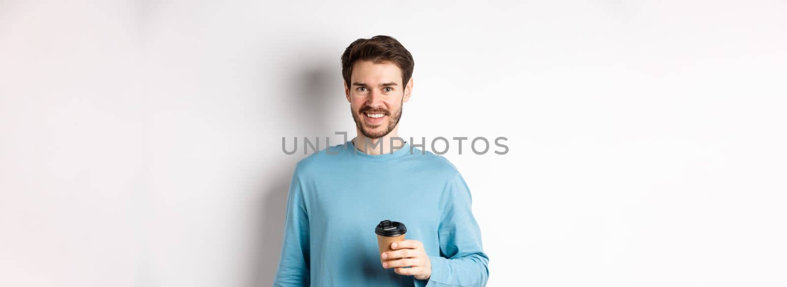 Cheerful man drinking coffee from cafe take-away and smiling, standing with paper cup in white background.