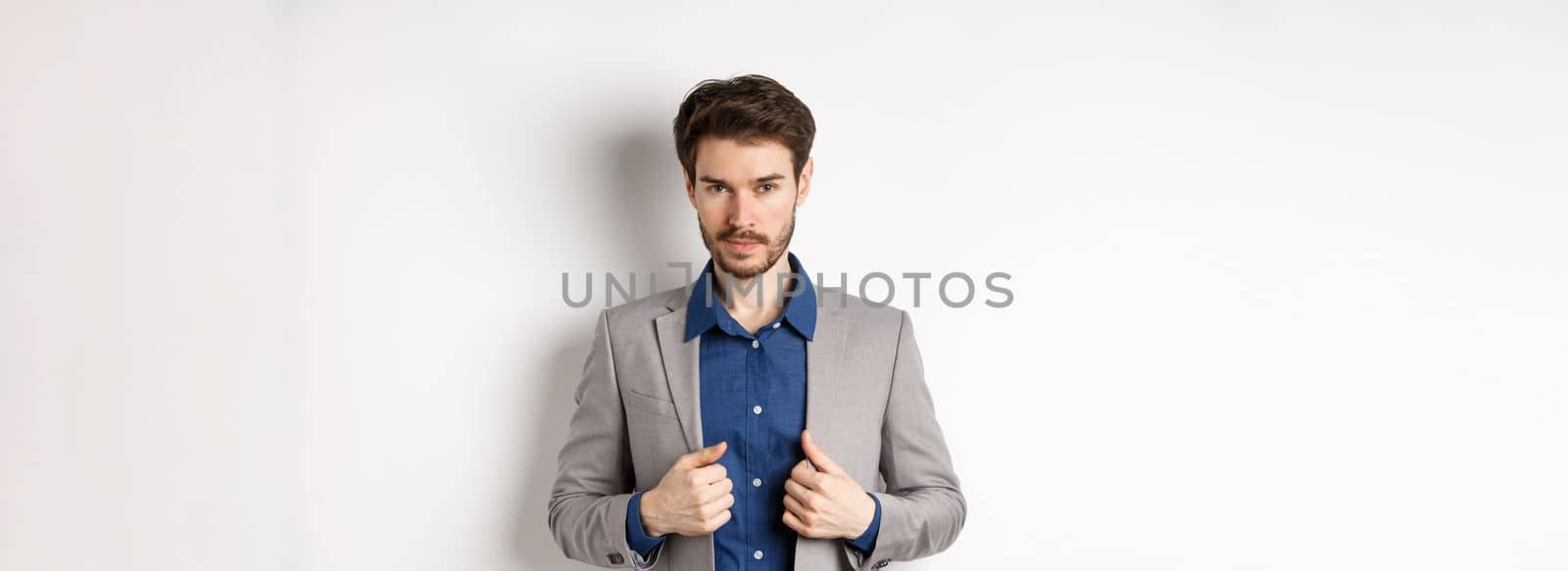 Confident young man with beard fixing suit and looking sassy at camera, feeling ready and determined, standing on white background.