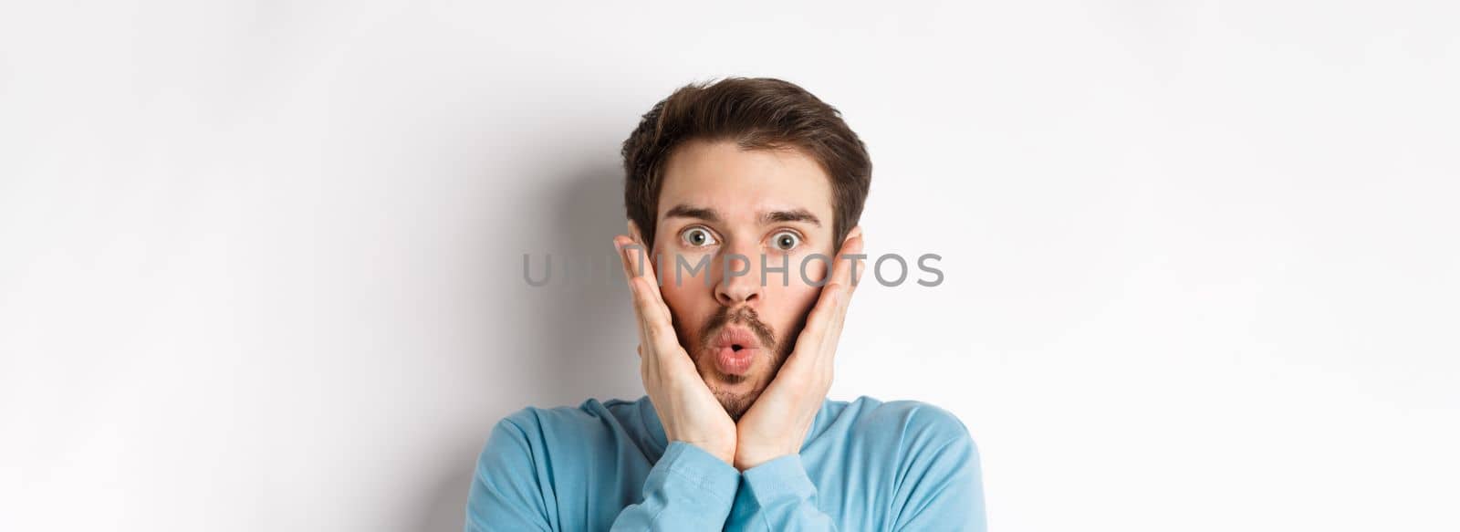 Close-up of impressed man checking out special offer, saying wow and touching face with amazed expression, stare at camera in awe, standing over white background.