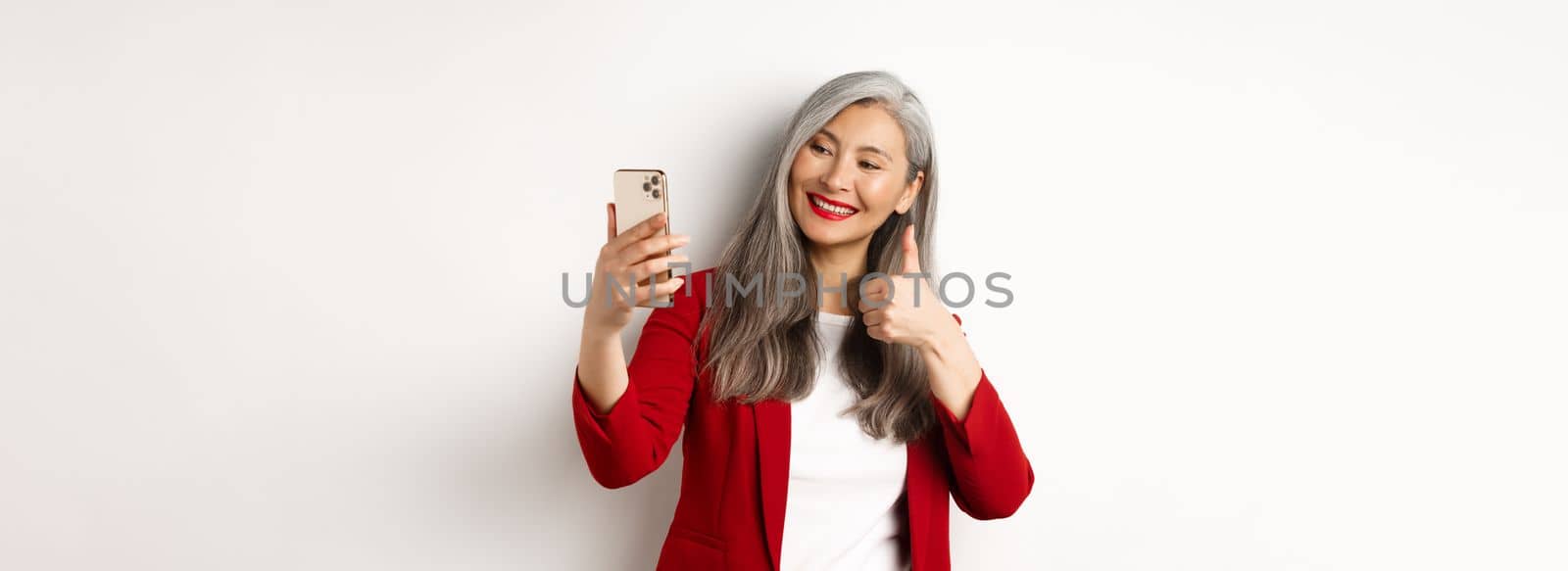 Successful asian businesswoman in red blazer, taking selfie on smartphone with thumb-up, showing approval, standing over white background.