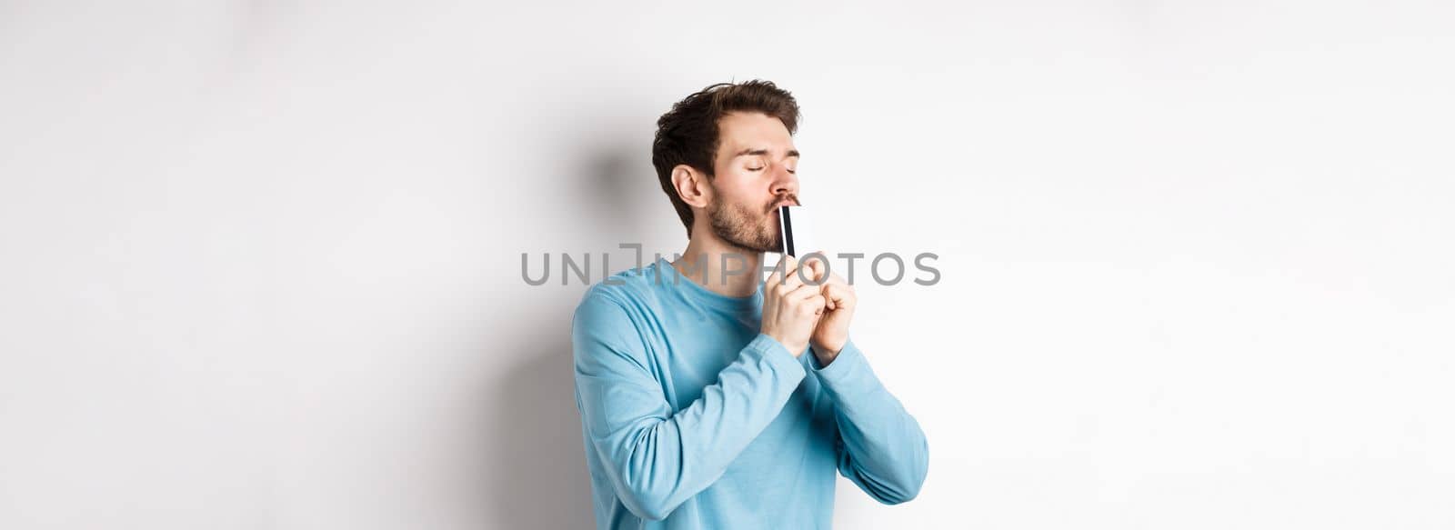 Young man kissing plastic credit card with satisfied face, standing over white background in blue casual shirt.