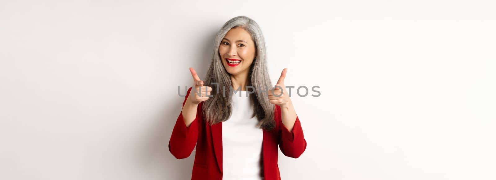 Professional female employer in trendy red blazer and makeup, pointing fingers at camera and smiling, praising something, need you, standing over white background.