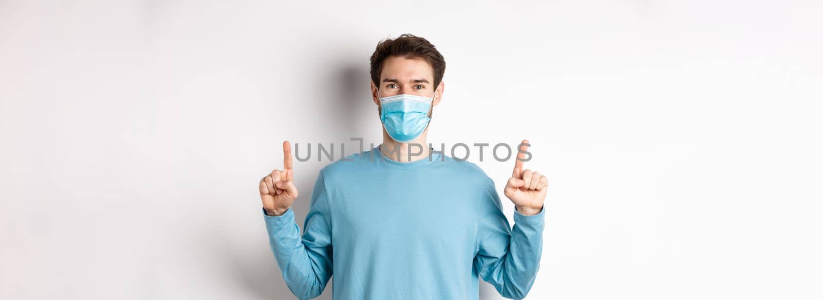 Coronavirus, health and quarantine concept. Handsome caucasian male model showing banner on top, pointing fingers up and wear medical mask, white background by Benzoix