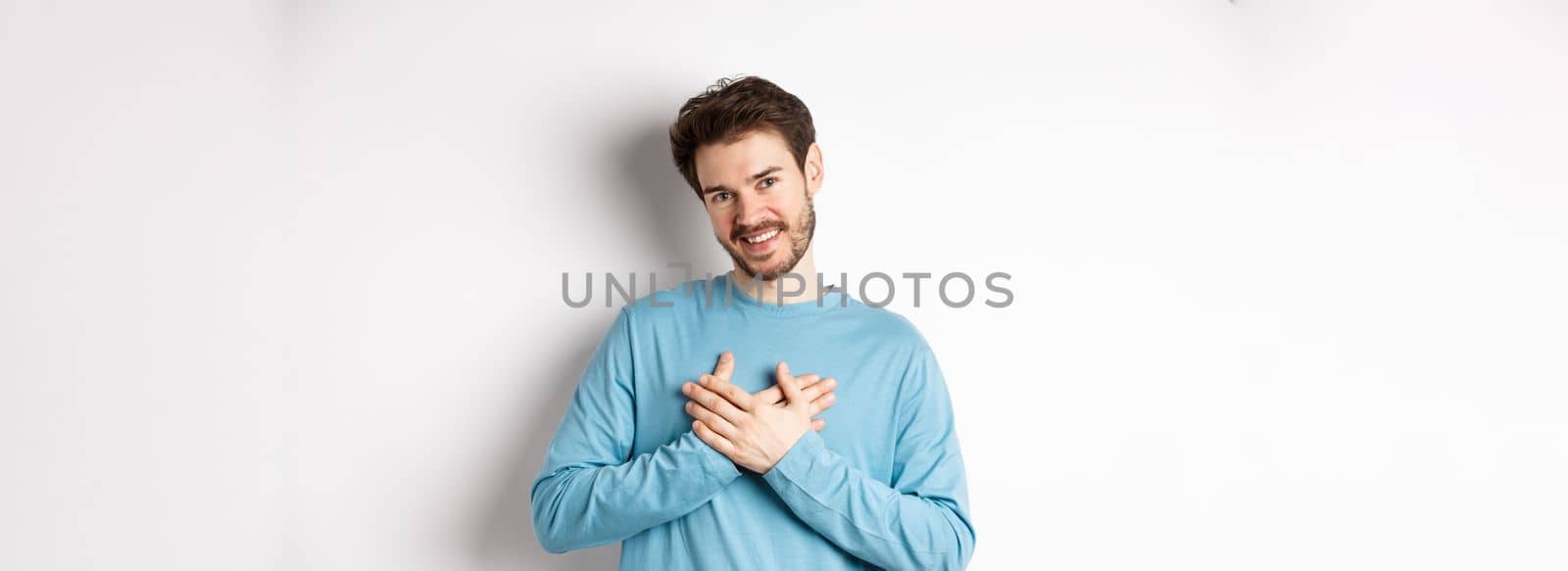 Portrait of young handsome man with beard, holding hands on heart and saying thank you, thinking about pleasant moment, standing over white background.