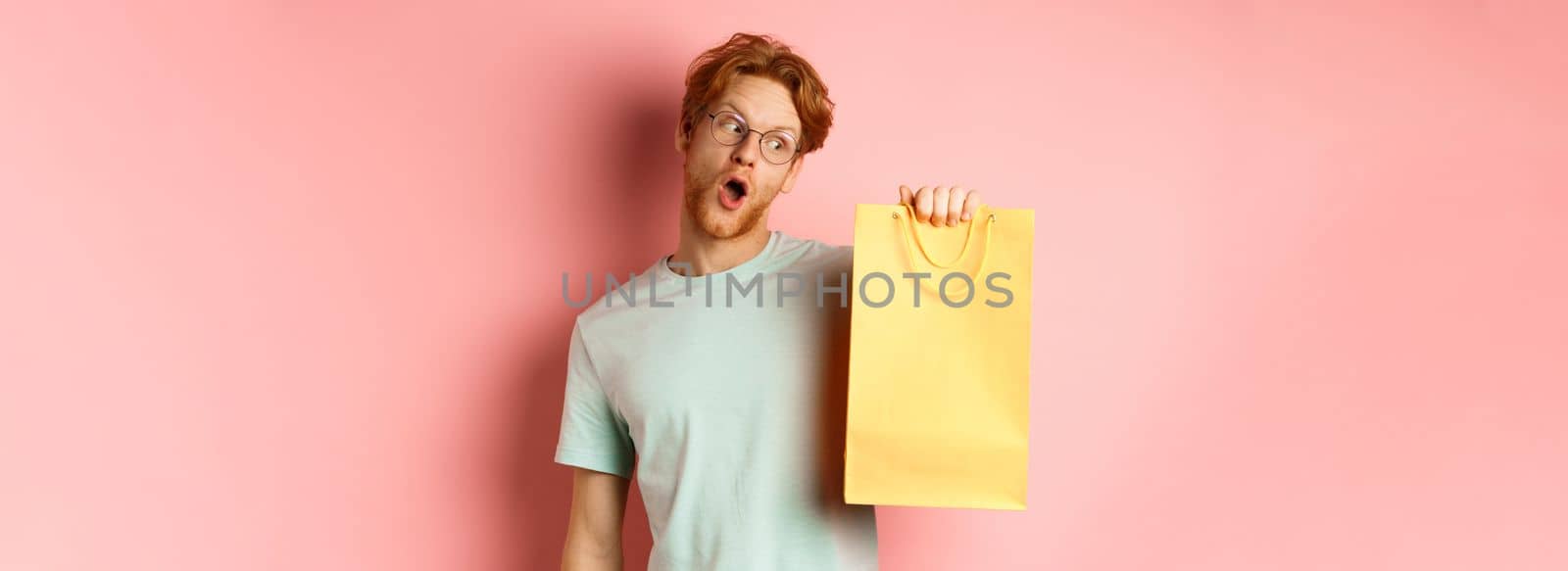 Funny handsome man with red hair, wearing glasses and t-shirt, holding and looking at yellow shopping bag, buying gifts during promo offer, standing over pink background.