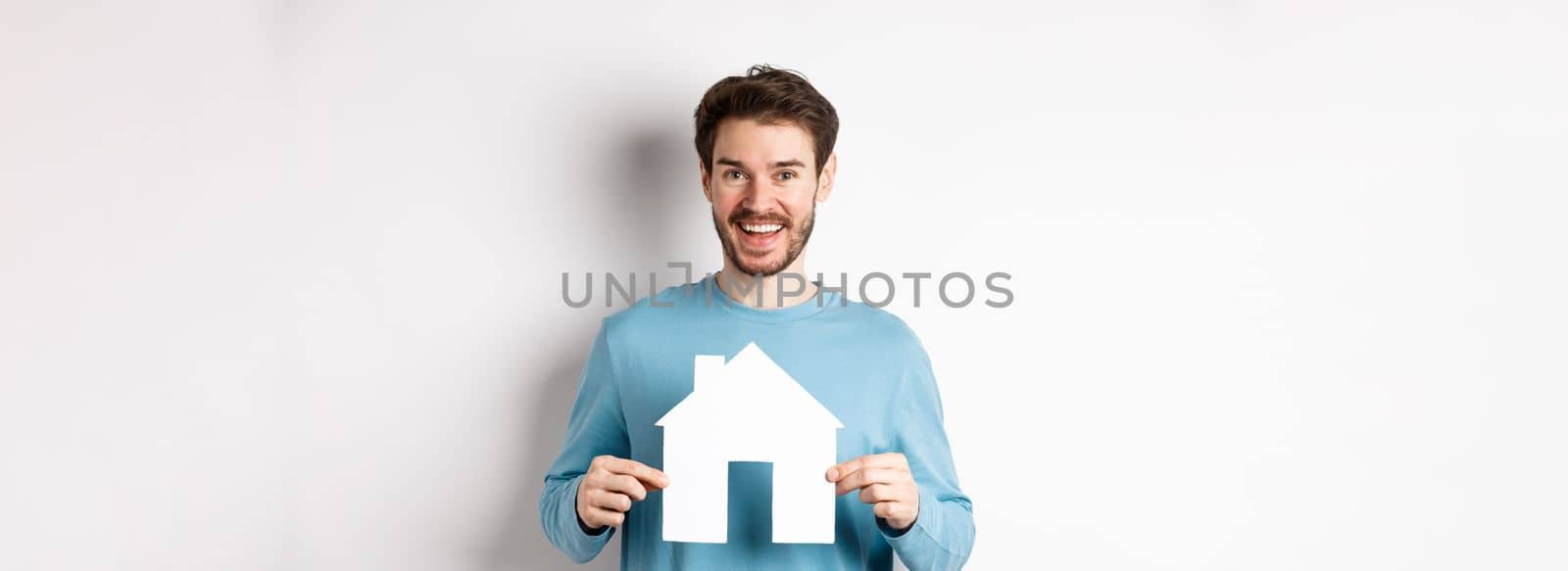 Real estate and insurance concept. Handsome modern man buying property, smiling and showing paper house cutout, standing over white background by Benzoix