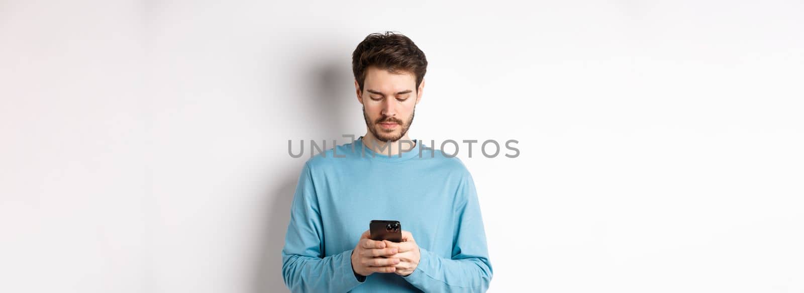 Serious young man reading message on smartphone, looking at mobile screen, standing over white background by Benzoix