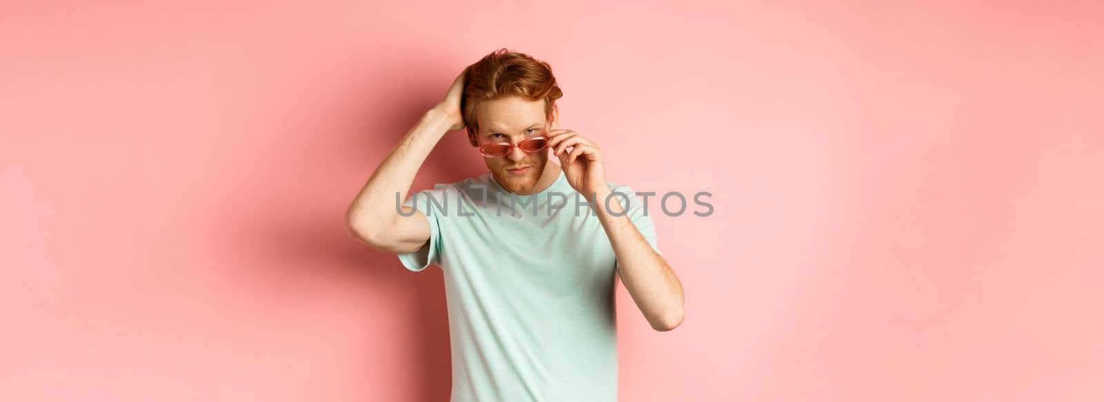 Handsome young redhead man in sunglasses, brushing hair with hand and looking smug and confident at camera, standing over pink background.