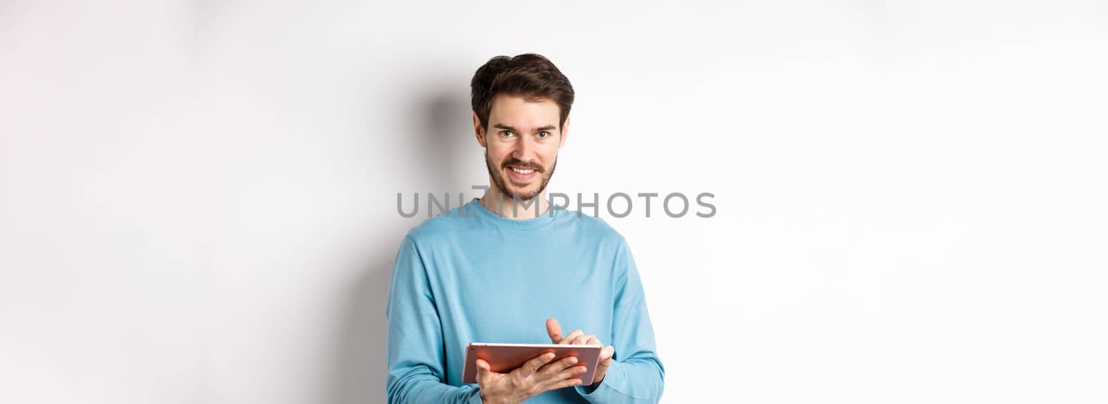 E-commerce. Smiling caucasian man using digital tablet and looking at camera, standing on white background.