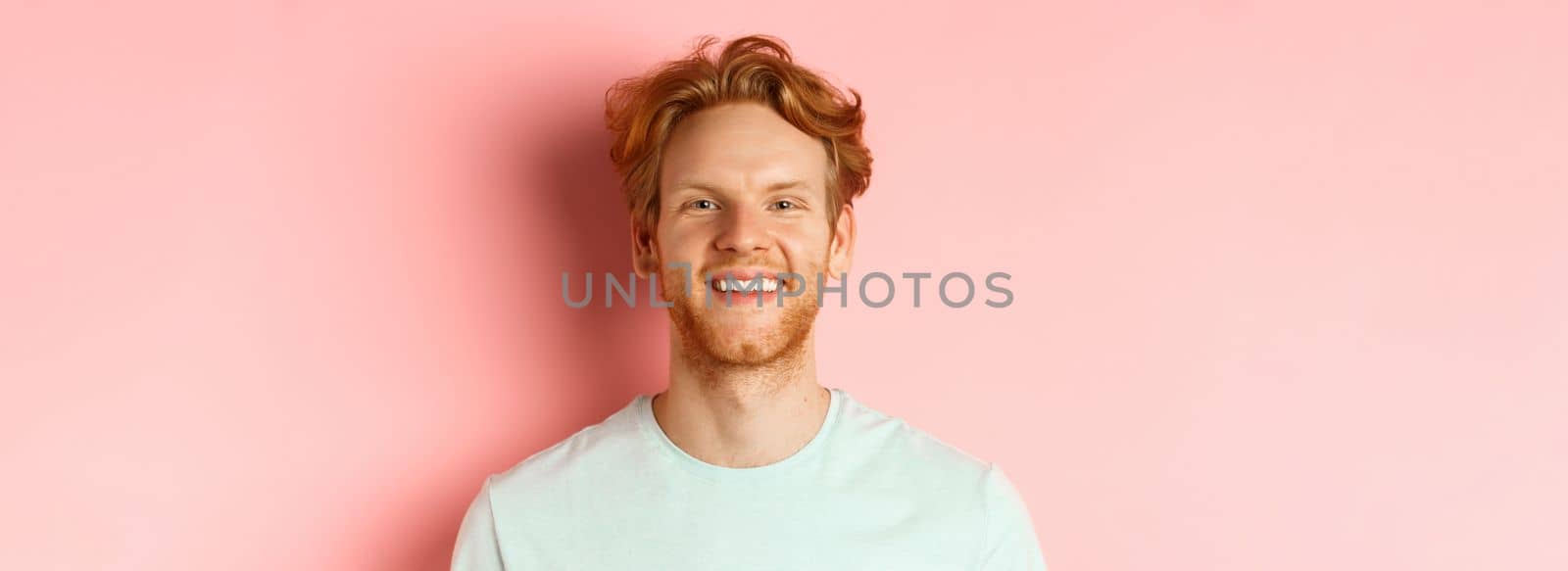 Happy young man with beard and messsy red haircut, smiling with white teeth and cheerful expression, standing over pink background.