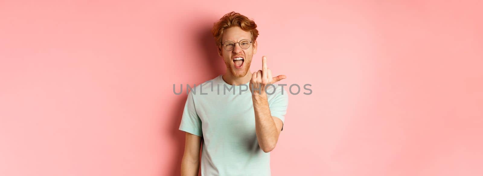 Arrogant and rude redhead man in glasses dont give a fuck, showing middle fingers at camera and frowning, standing over pink background.