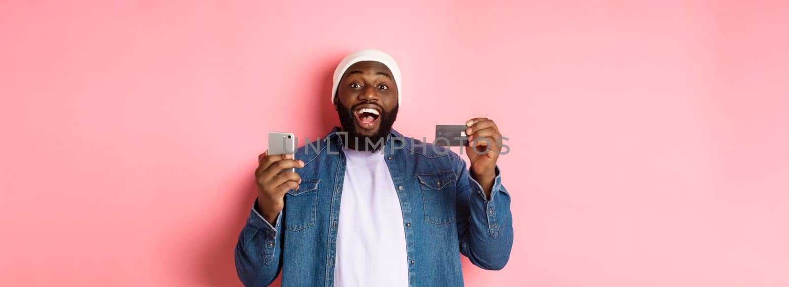 Online shopping. Happy bearded african-american man smiling, showing credit card and making purchase on smartphone, standing over pink background.