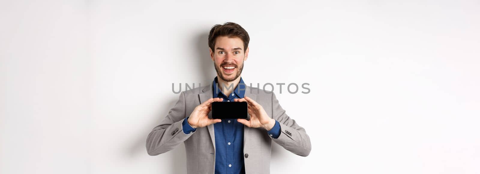 E-commerce and online shopping concept. Excited young businessman in suit showing empty smartphone, screen demonstrate app achievement, white background.