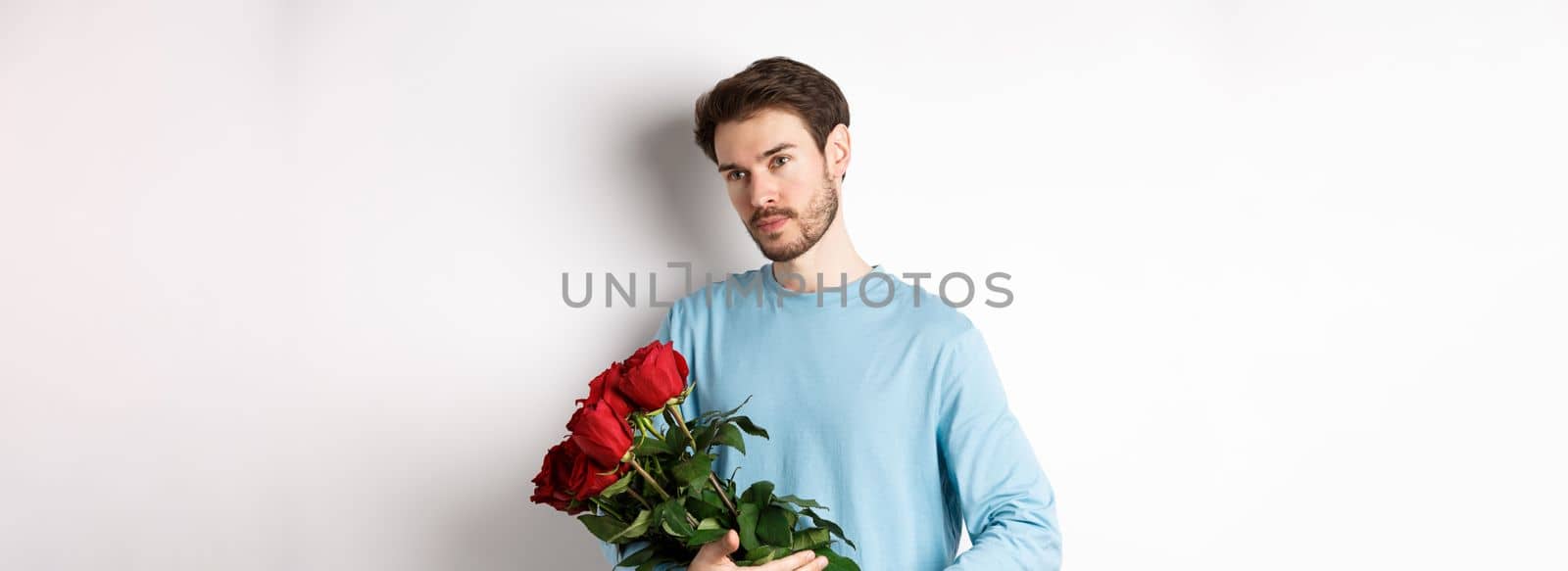 Handsome young man holding beautiful red roses for his lover on Valentines day, looking pensive, standing over white background.