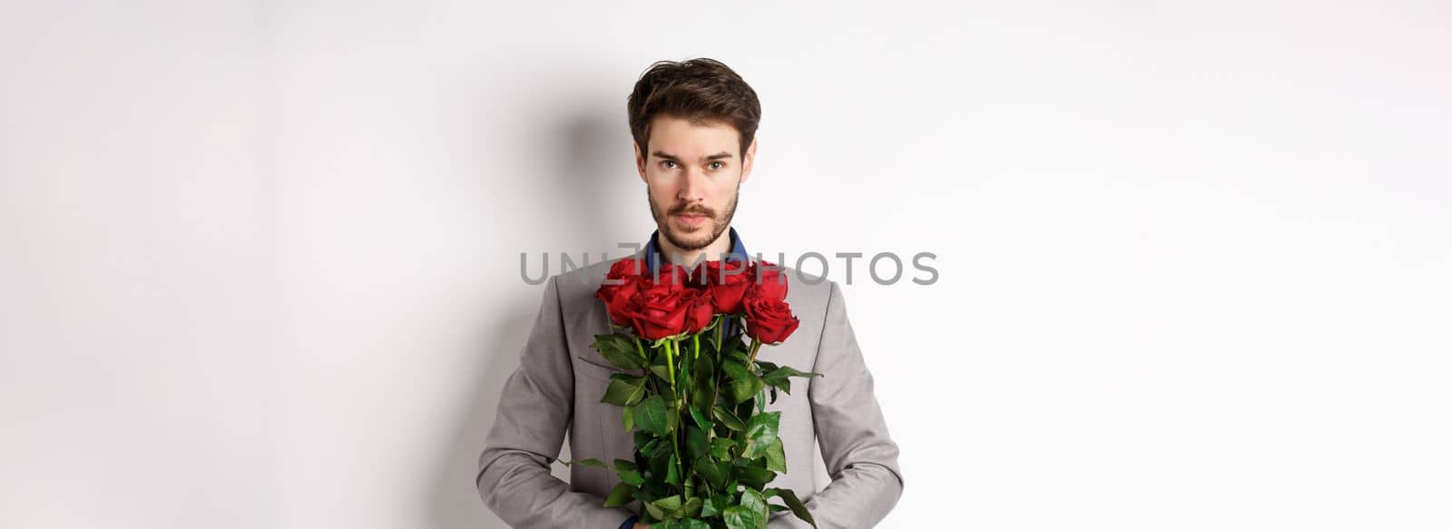 Valentines day and love concept. Confident and handsome man in suit holding bouquet of roses, looking at camera, standing over white background.