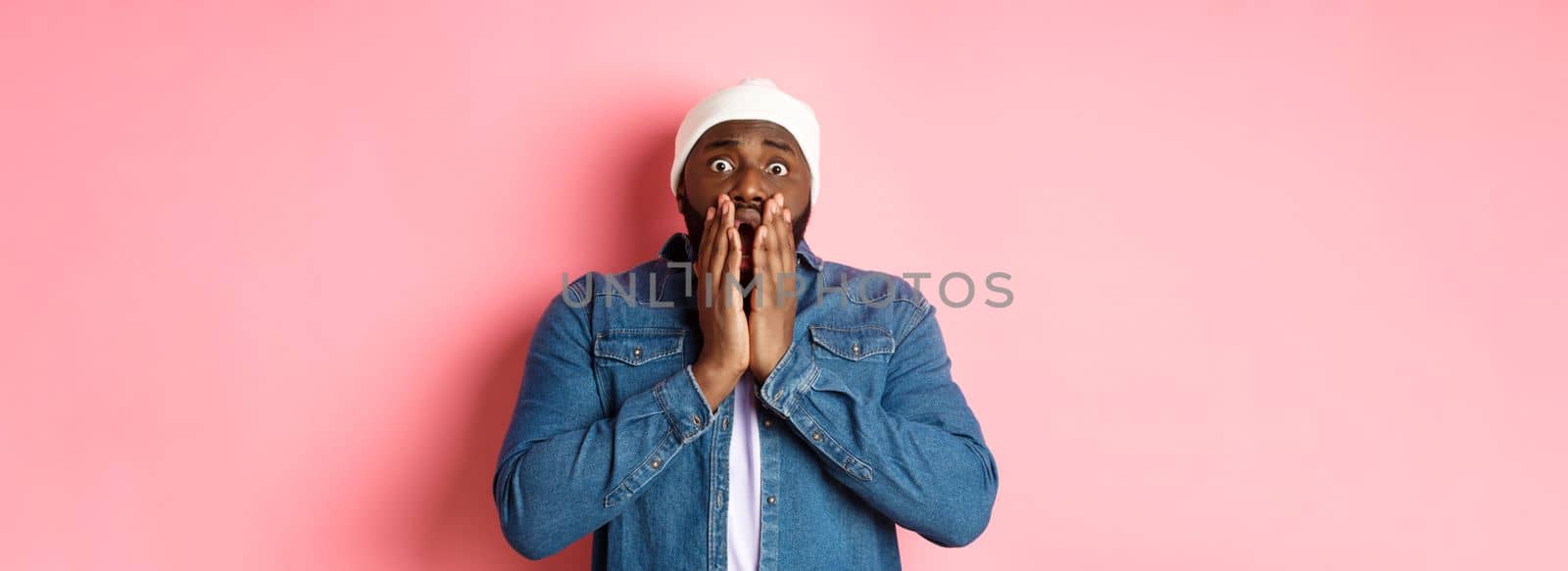 Alarmed and worried Black man staring at bad accident, gasping and standing in panic over pink background.