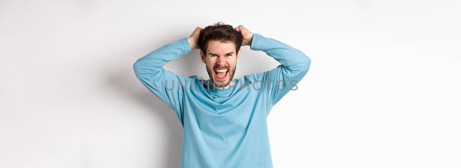 Mad young man pull hair oyt of head and screaming angry, shouting frustrated, standing hateful against white background.