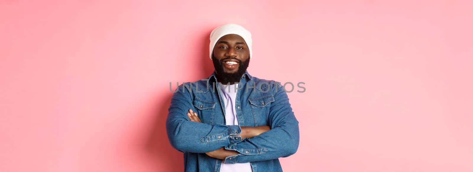 Handsome hip hop style Black man in beanie and denim shirt, smiling confident, cross arms on chest and staring at camera on pink background.