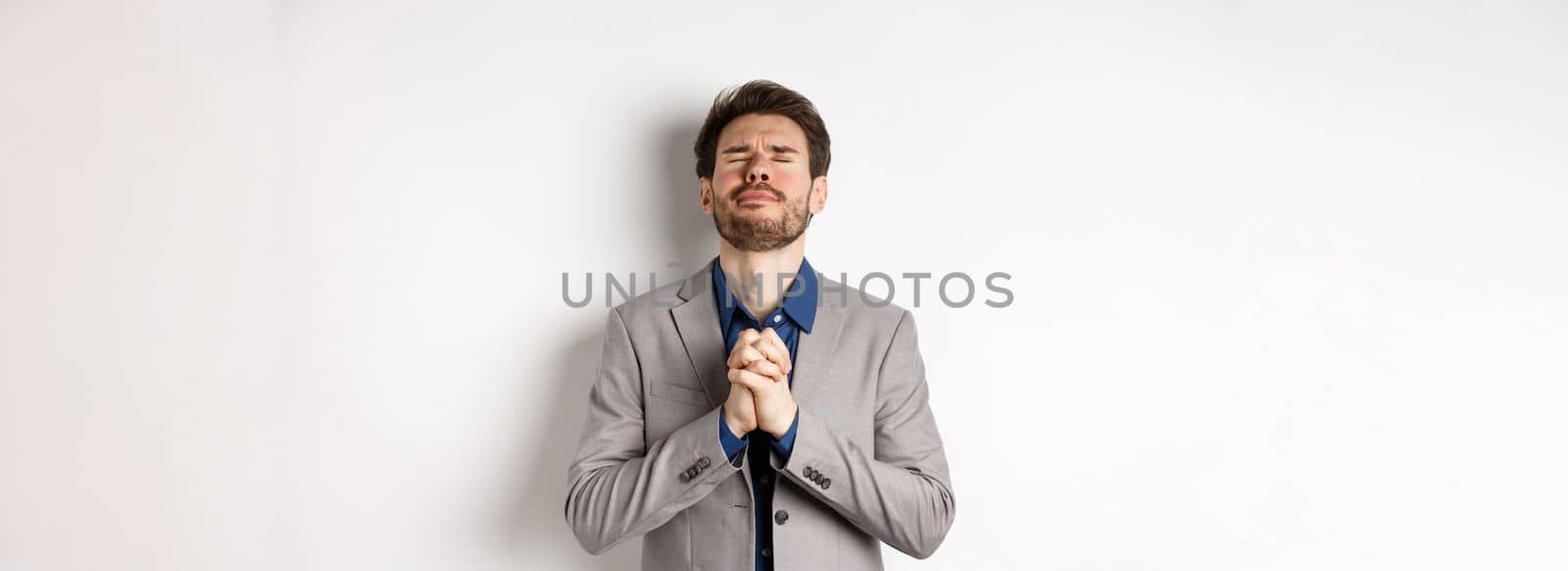 Nervous male entrepreneur in suit begging god, holding hands in pray with pleading expression, close eyes and make wish, asking for help, standing on white background by Benzoix