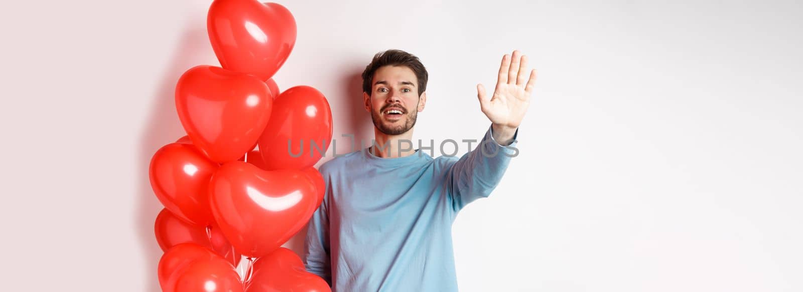 Handsome young caucasian man standing with romantic heart balloon and waving hand at lover, waiting for his date on Valentines day with cute surprise, standing over white background.