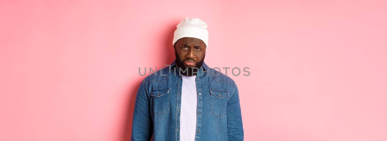 Disappointed and sad Black man, sulking and whining, looking at camera with offended grimace, standing over pink background.
