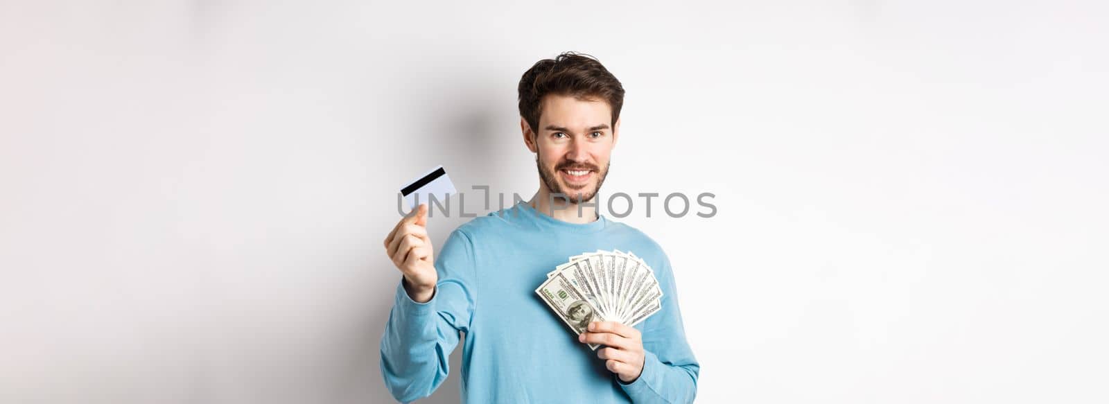 Handsome young man smiling and offering payment in cash and contactless, showing money with plastic credit card, standing on white background by Benzoix