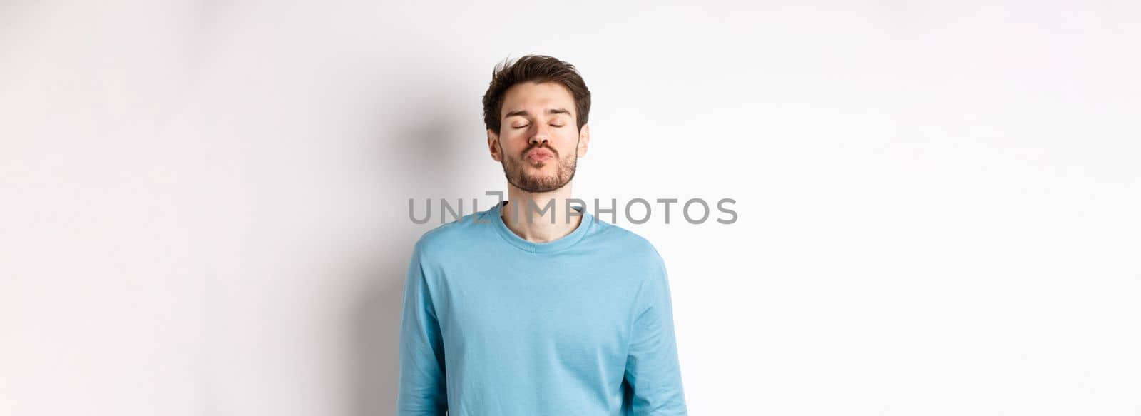 Attractive boyfriend waiting for kiss with puckered lips and closed eyes, standing in sweatshirt over white background.