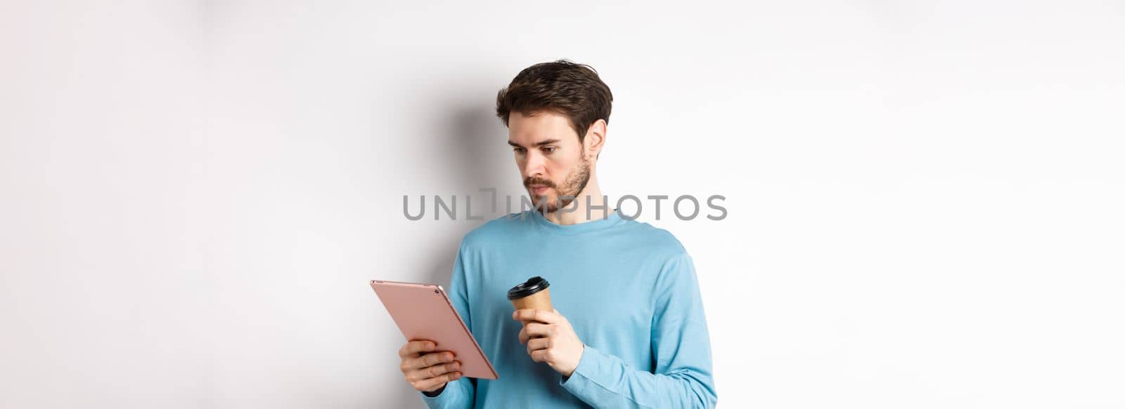 Man reading news online in digital tablet, looking serious at screen while drinking coffee from paper cup, standing over white background.